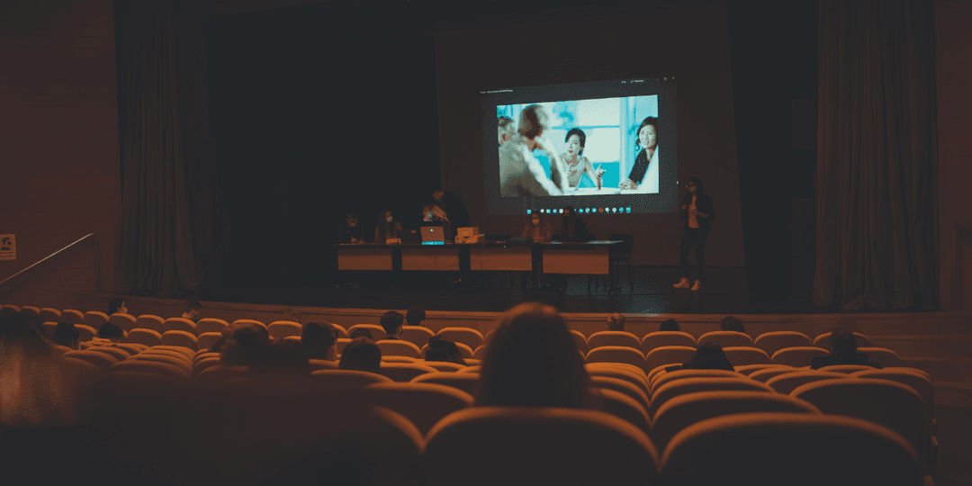 A presentation scene with speakers on stage and an audience in a dark auditorium
