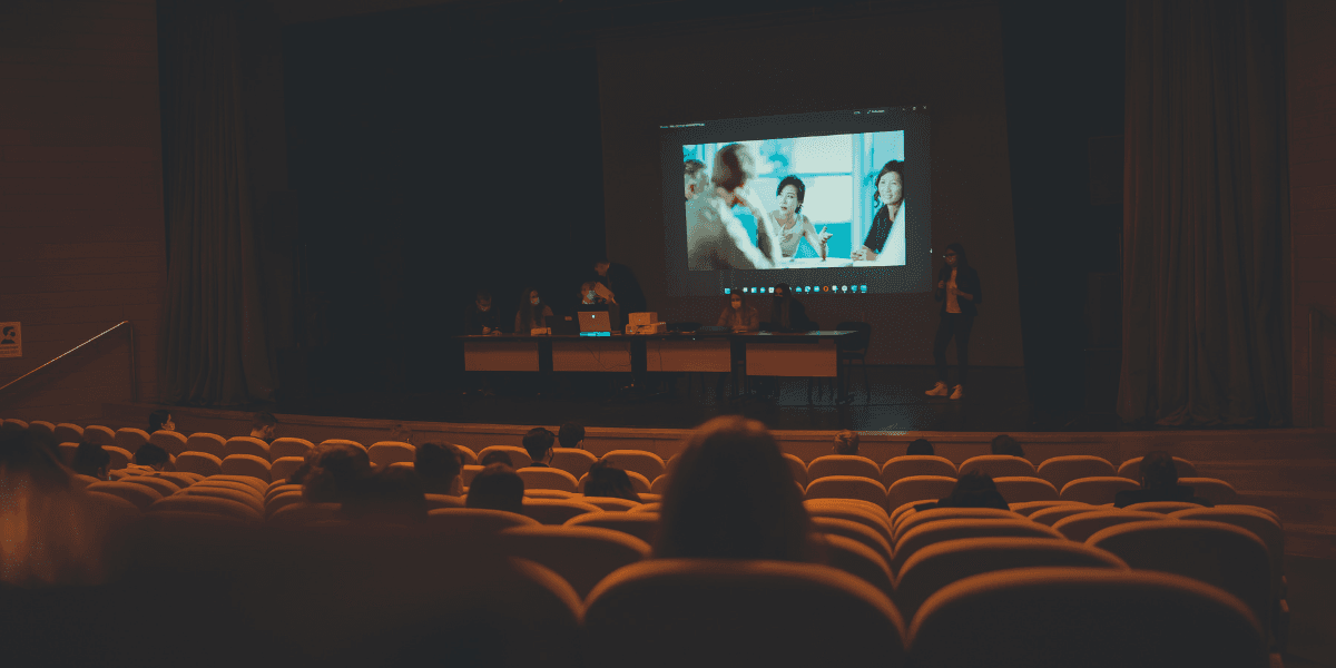 A presentation scene with speakers on stage and an audience in a dark auditorium