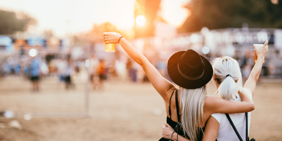 Two friends enjoying a festival sunset, raising their drinks while embracing each other.
