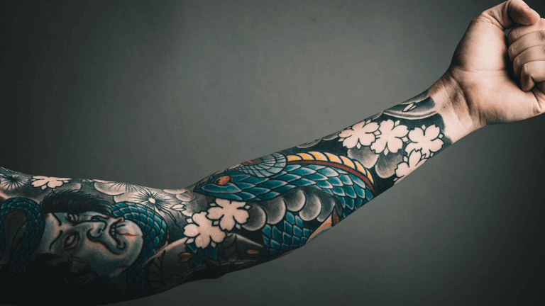 Colorful Japanese-style tattoo sleeve featuring a serpent, flowers, and portrait on an outstretched arm.