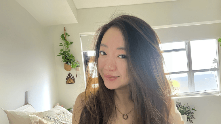 Daphne Ooi, a peaceful Asian woman with long hair, eyes closed, meditates in a serene bedroom filled with plants.