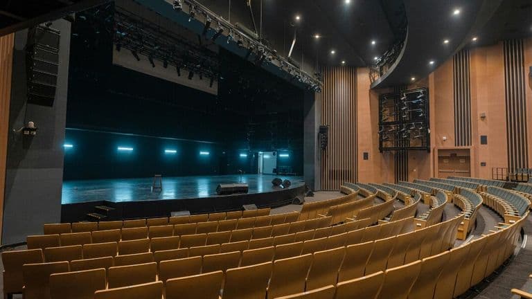 A spacious, modern theater with curved rows of empty yellow seats facing a brightly lit stage. The atmosphere is calm and anticipatory.