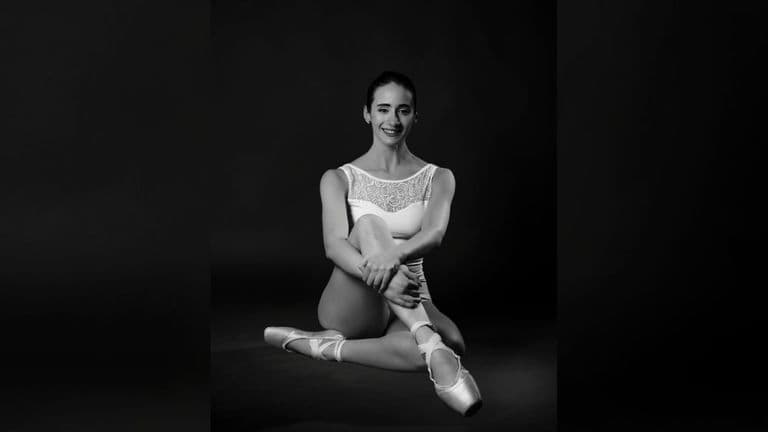 A dancer in a white leotard and pointe shoes sits gracefully, exuding elegance, captured in black and white.