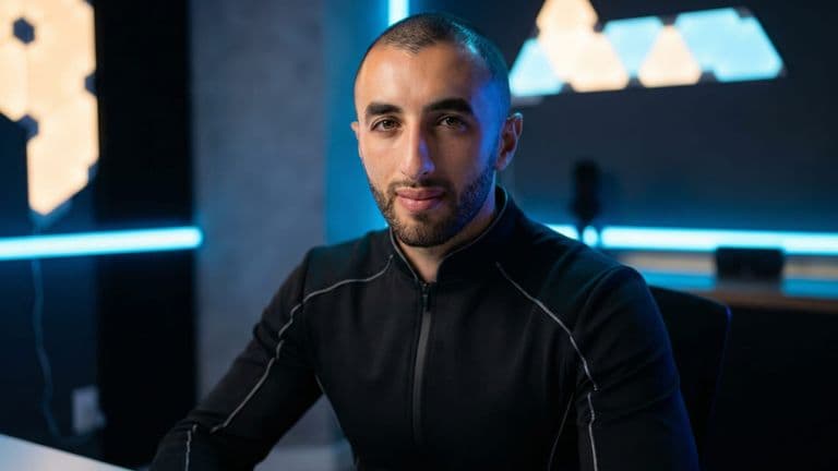 Portrait of a man in a modern, blue-lit workspace wearing a black zip-up jacket and looking at the camera.