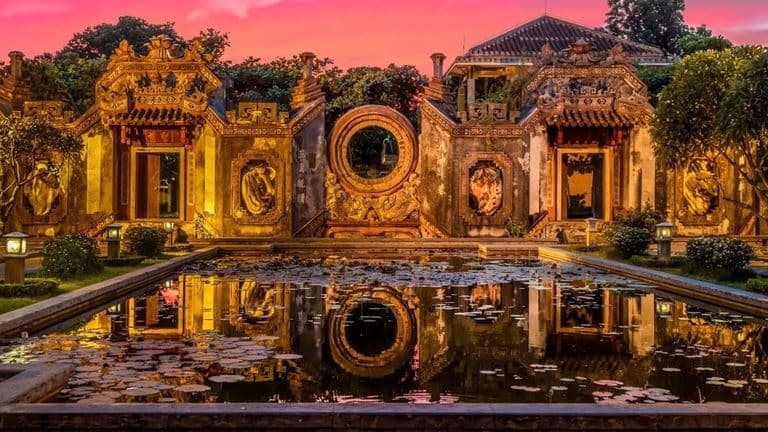 Ornate Vietnamese architectural structure at dusk, reflecting in a tranquil pond surrounded by lush greenery.