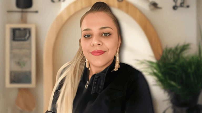 Portrait of a woman with long, blonde hair and golden earrings, wearing a black jacket, smiling confidently in front of a neutral background with plants