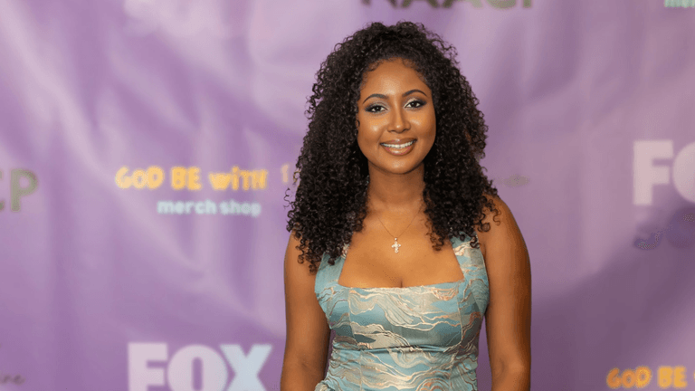 Smiling young woman with curly hair, wearing a light blue dress, standing in front of a purple backdrop with logos for the NAACP and FOX. She has a cross necklace and a radiant expression.
