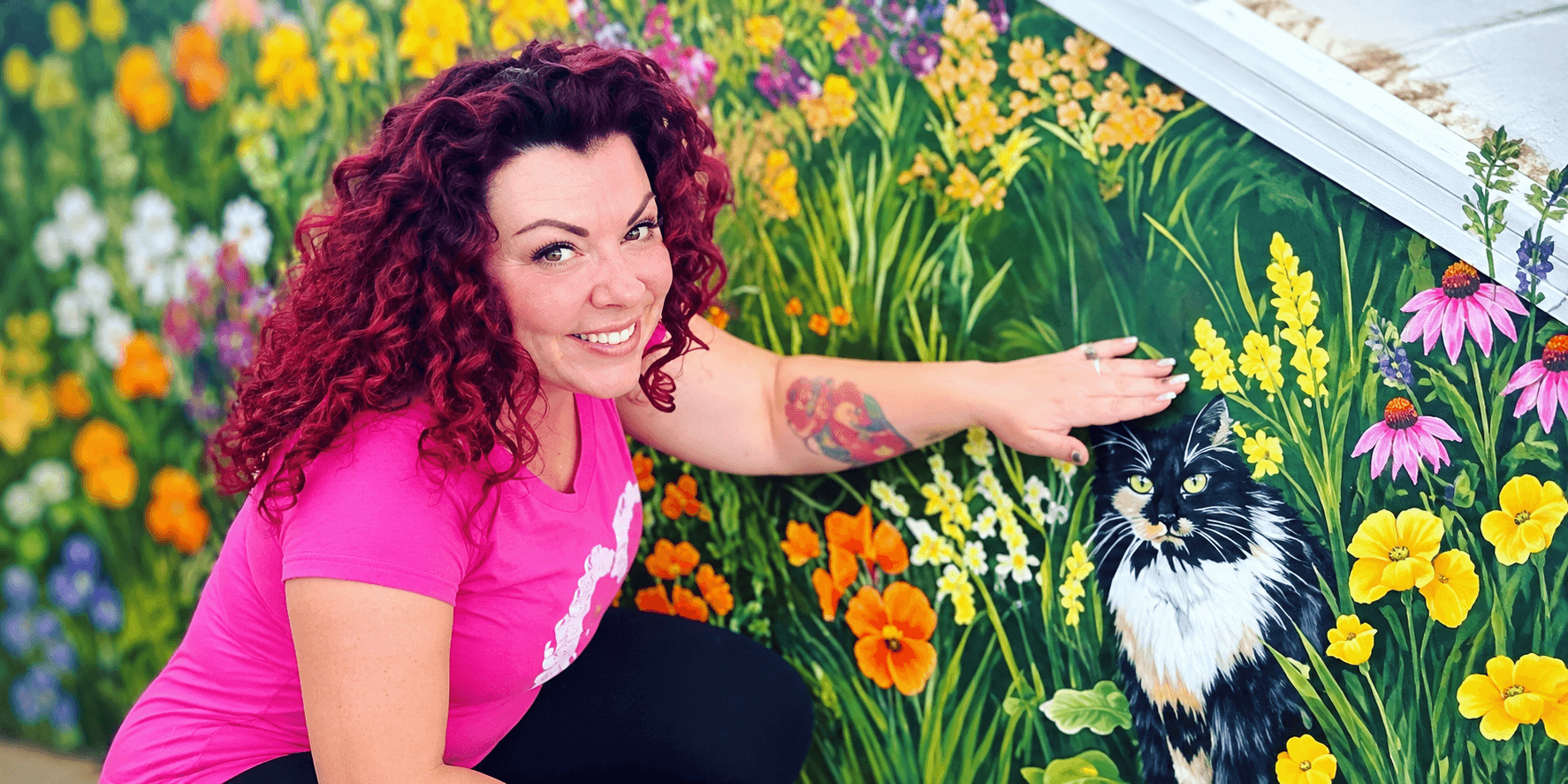 Woman with curly red hair poses by a vibrant mural featuring flowers and a cat