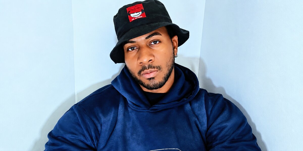 Portrait of Ju-Lion The Voice wearing a black bucket hat and navy hoodie against a light blue background.