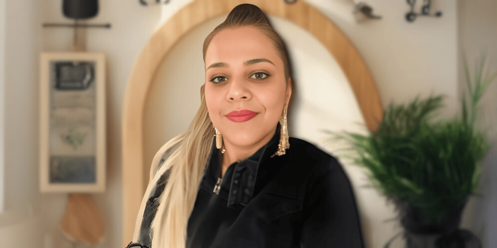 Portrait of a woman with long, blonde hair and golden earrings, wearing a black jacket, smiling confidently in front of a neutral background with plants