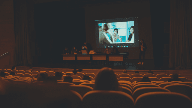 A presentation scene with speakers on stage and an audience in a dark auditorium