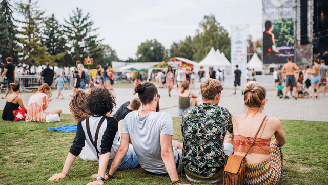A group of people sitting on grass, enjoying a festival atmosphere with a stage nearby