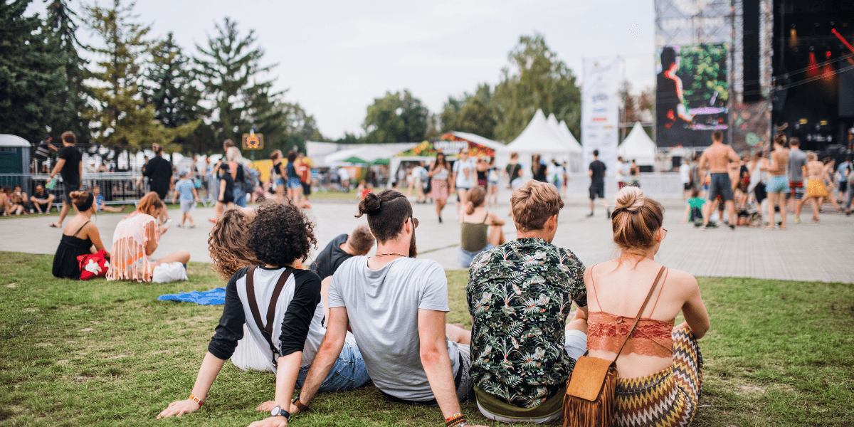 A group of people sitting on grass, enjoying a festival atmosphere with a stage nearby
