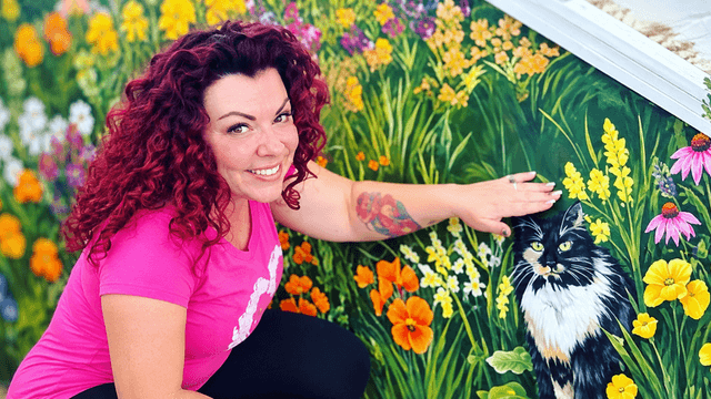 Woman with curly red hair poses by a vibrant mural featuring flowers and a cat