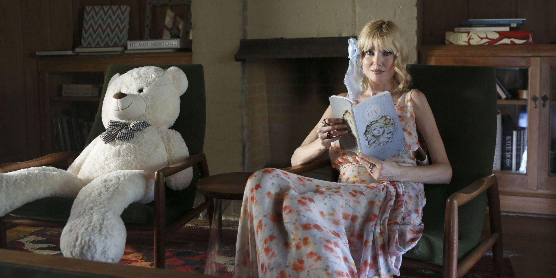 A woman in a floral dress reads a book beside a large teddy bear.