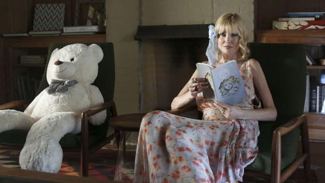 A woman in a floral dress reads a book beside a large teddy bear.