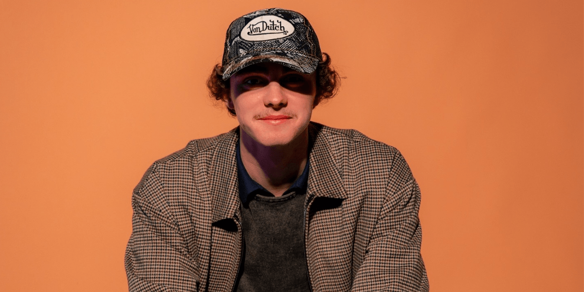 A portrait of Blake Benson, a young music industry innovator and the founder of Project A&R, showcasing his stylish, relaxed attire and confident demeanor against a vibrant orange background, symbolizing his fresh approach to artist representation and music marketing.