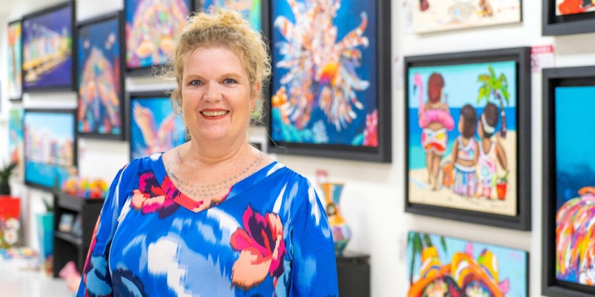 Portrait of a smiling artist standing in front of vibrant artwork featuring bright tropical designs and abstract art.