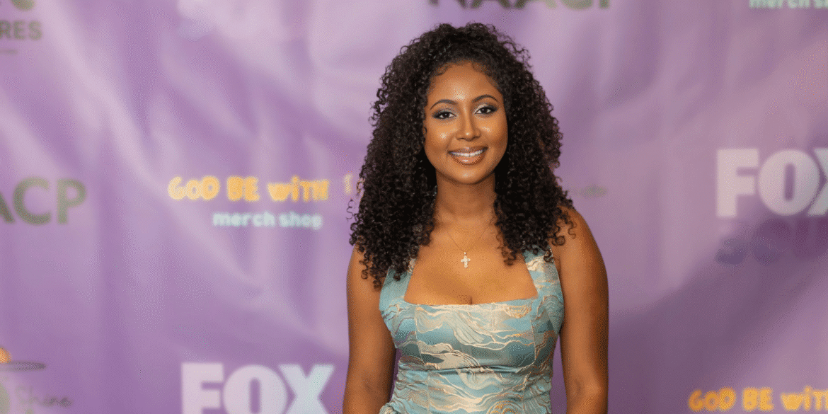 Smiling young woman with curly hair, wearing a light blue dress, standing in front of a purple backdrop with logos for the NAACP and FOX. She has a cross necklace and a radiant expression.