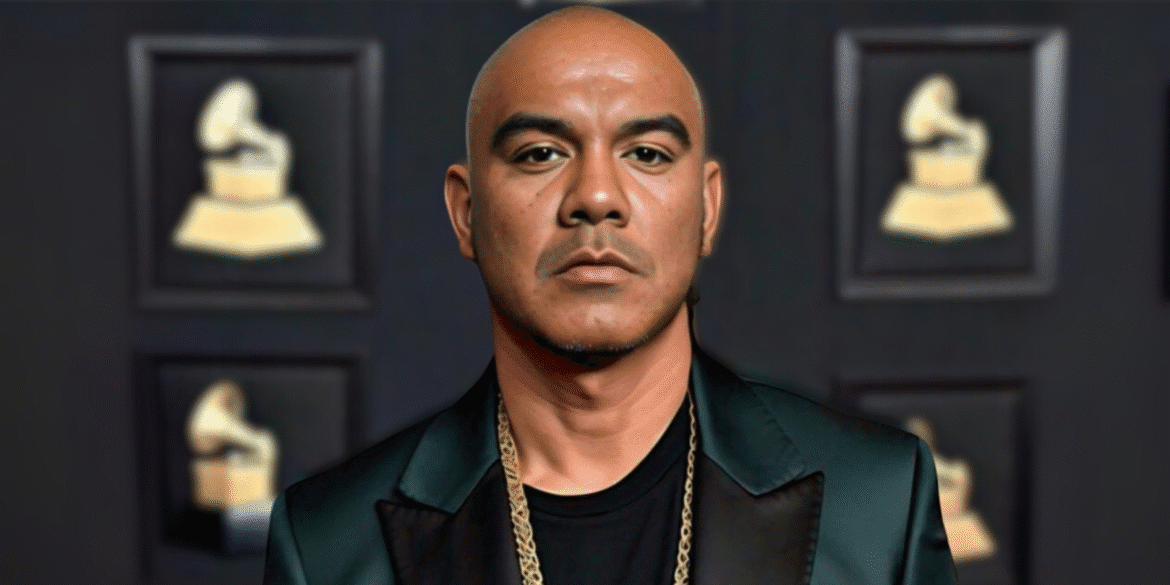 "RJ Kaneao, a Grammy-winning artist, standing confidently at a red carpet event, dressed in a black suit with a chain necklace."