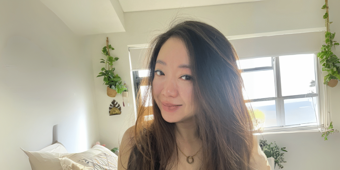 Daphne Ooi, a peaceful Asian woman with long hair, eyes closed, meditates in a serene bedroom filled with plants.