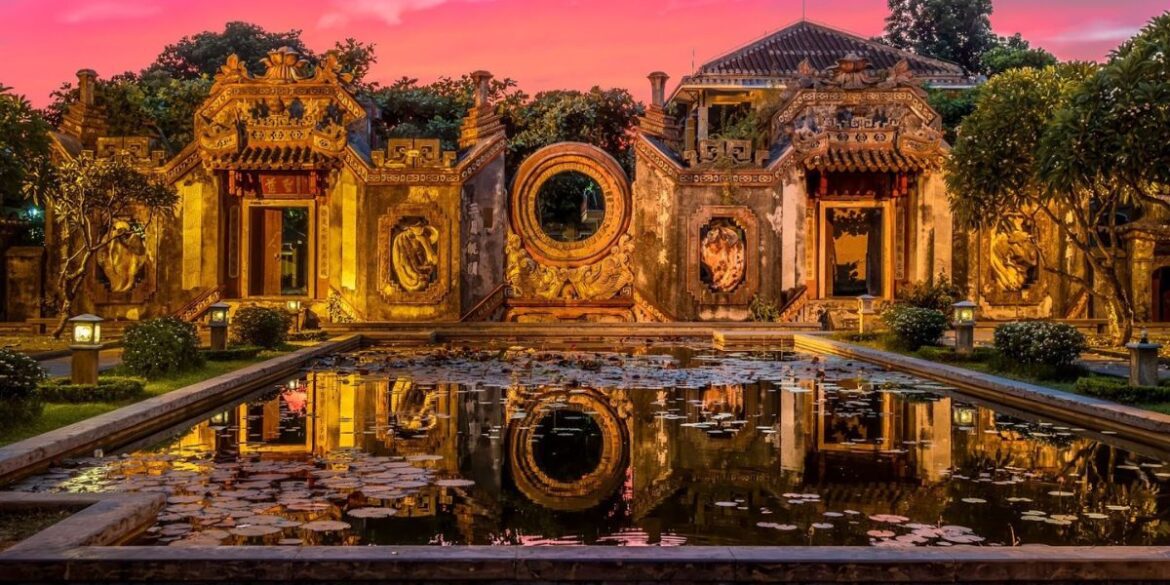 Ornate Vietnamese architectural structure at dusk, reflecting in a tranquil pond surrounded by lush greenery.