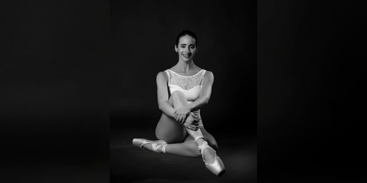 A dancer in a white leotard and pointe shoes sits gracefully, exuding elegance, captured in black and white.