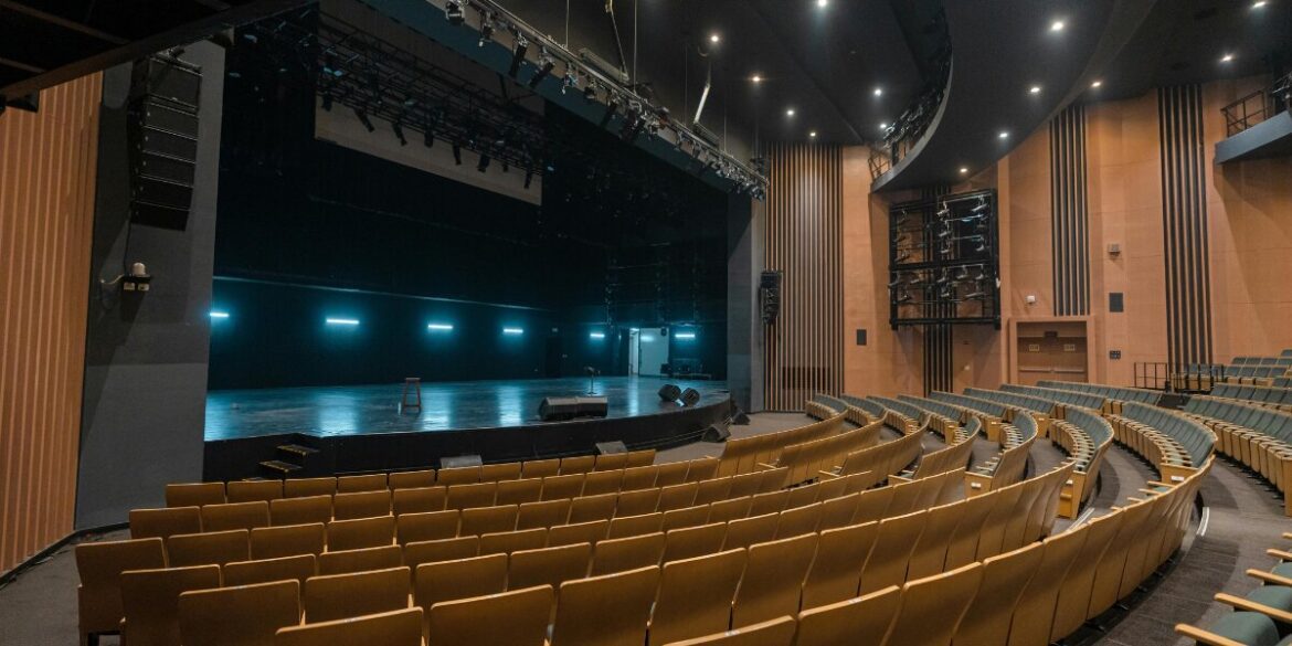 A spacious, modern theater with curved rows of empty yellow seats facing a brightly lit stage. The atmosphere is calm and anticipatory.