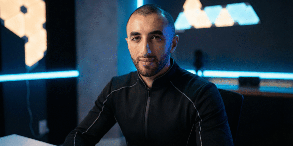 Portrait of a man in a modern, blue-lit workspace wearing a black zip-up jacket and looking at the camera.